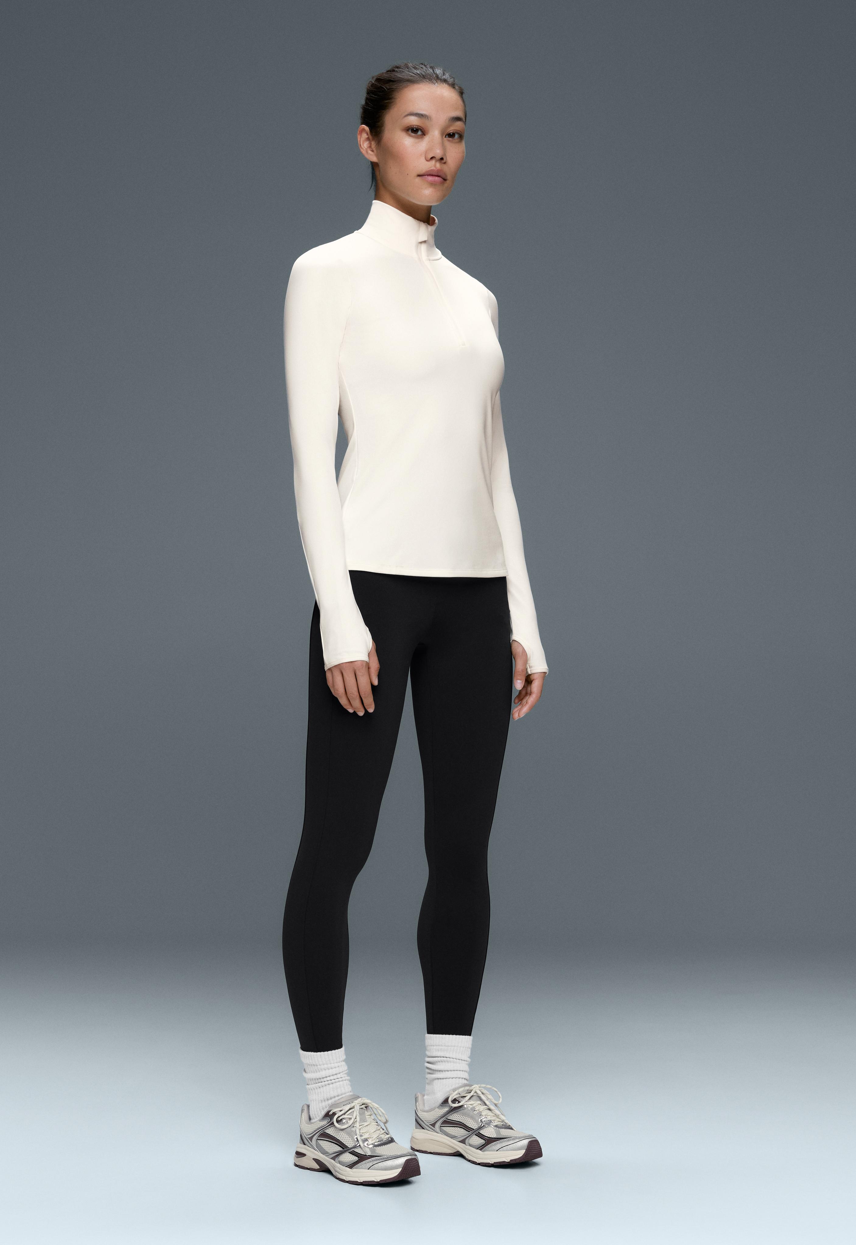 Fitted white long-sleeve top with a mock neck, half-zip closure and thumbholes, black leggings, and sneakers.