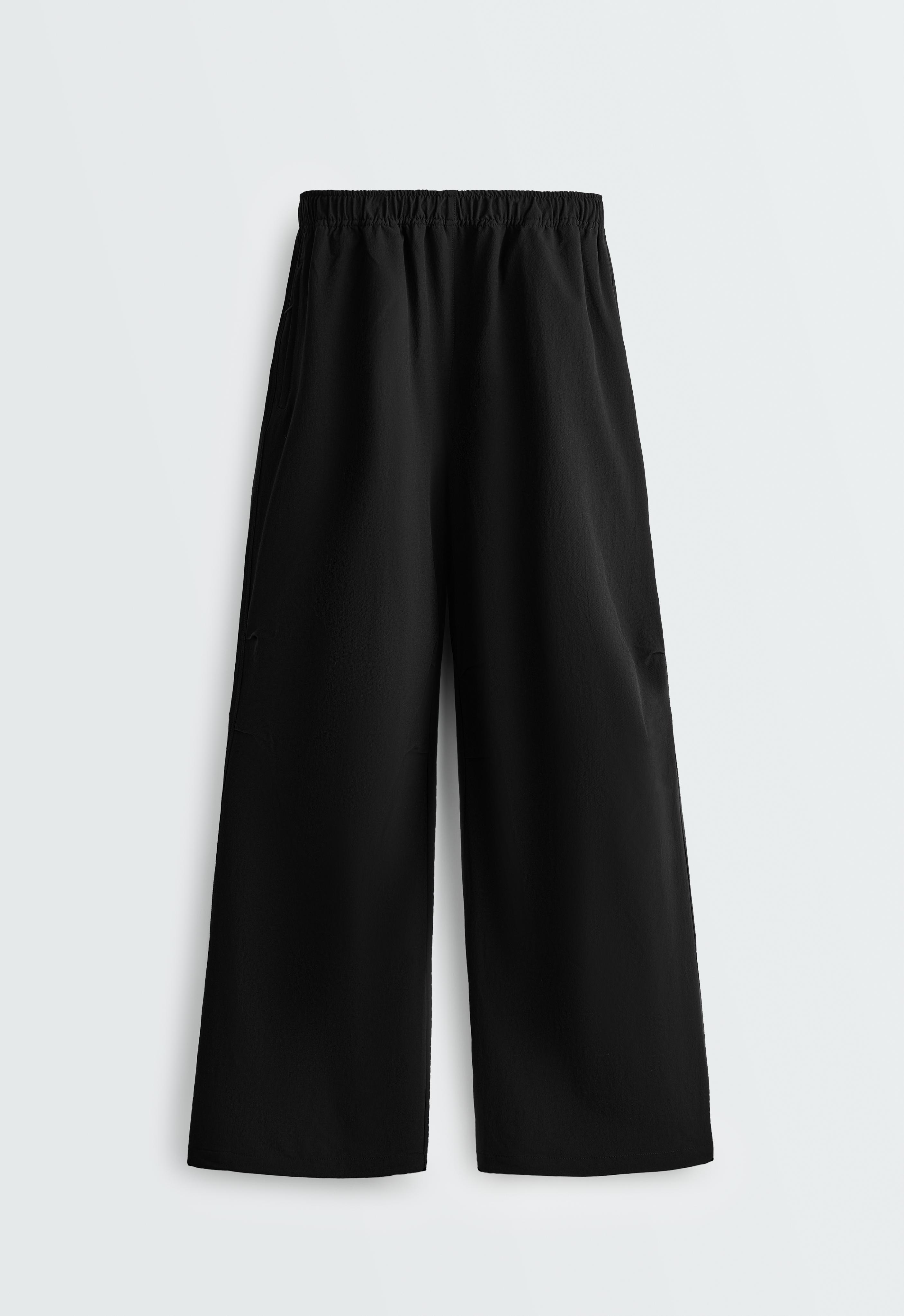 Front view of black wide-leg trousers with a gathered elastic waistband and pleated detail on the sides of the knees.