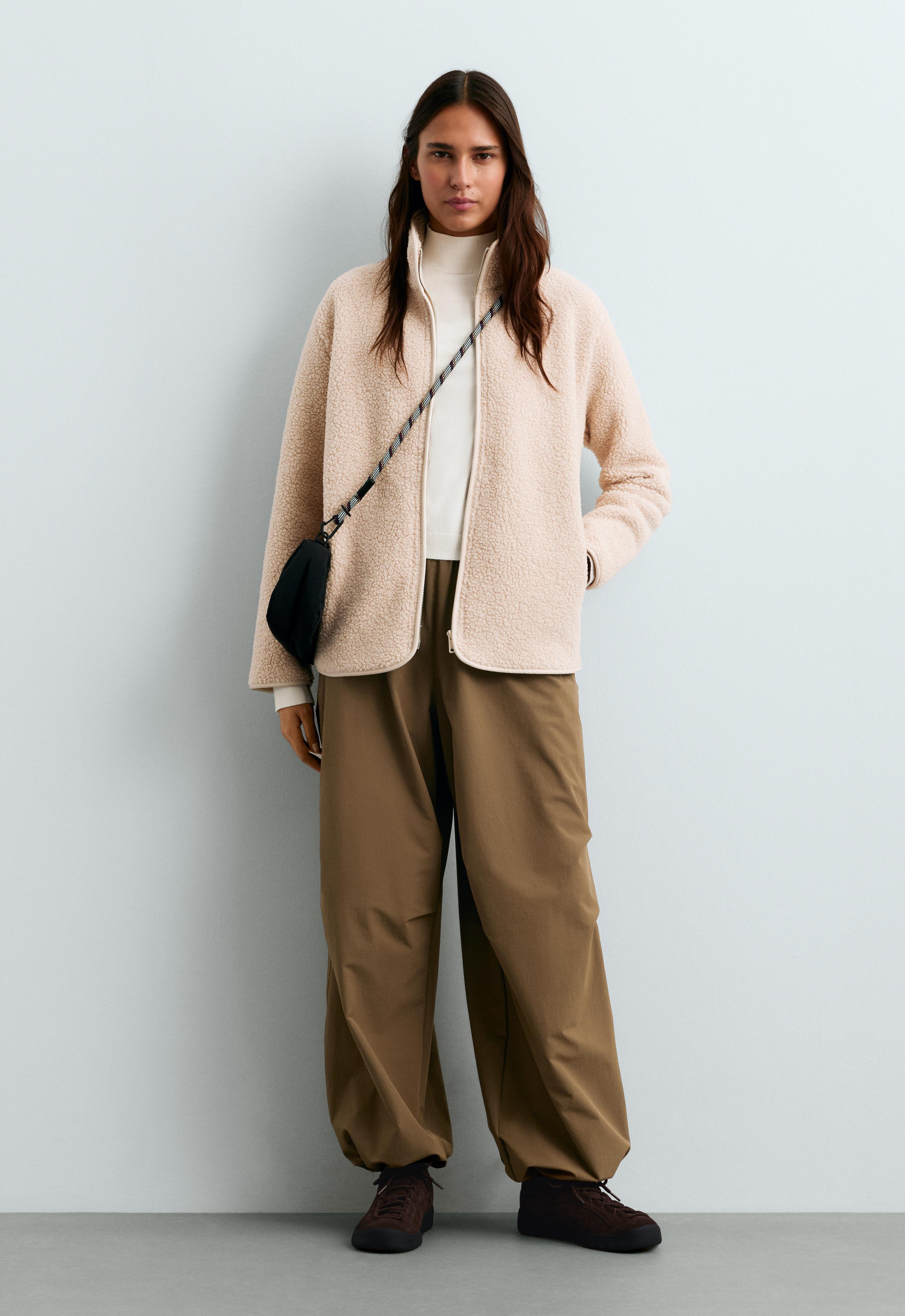 Cream-colored jacket set with a high collar, zipper, and side pockets, white top, brown pants, sneakers, and black handbag.