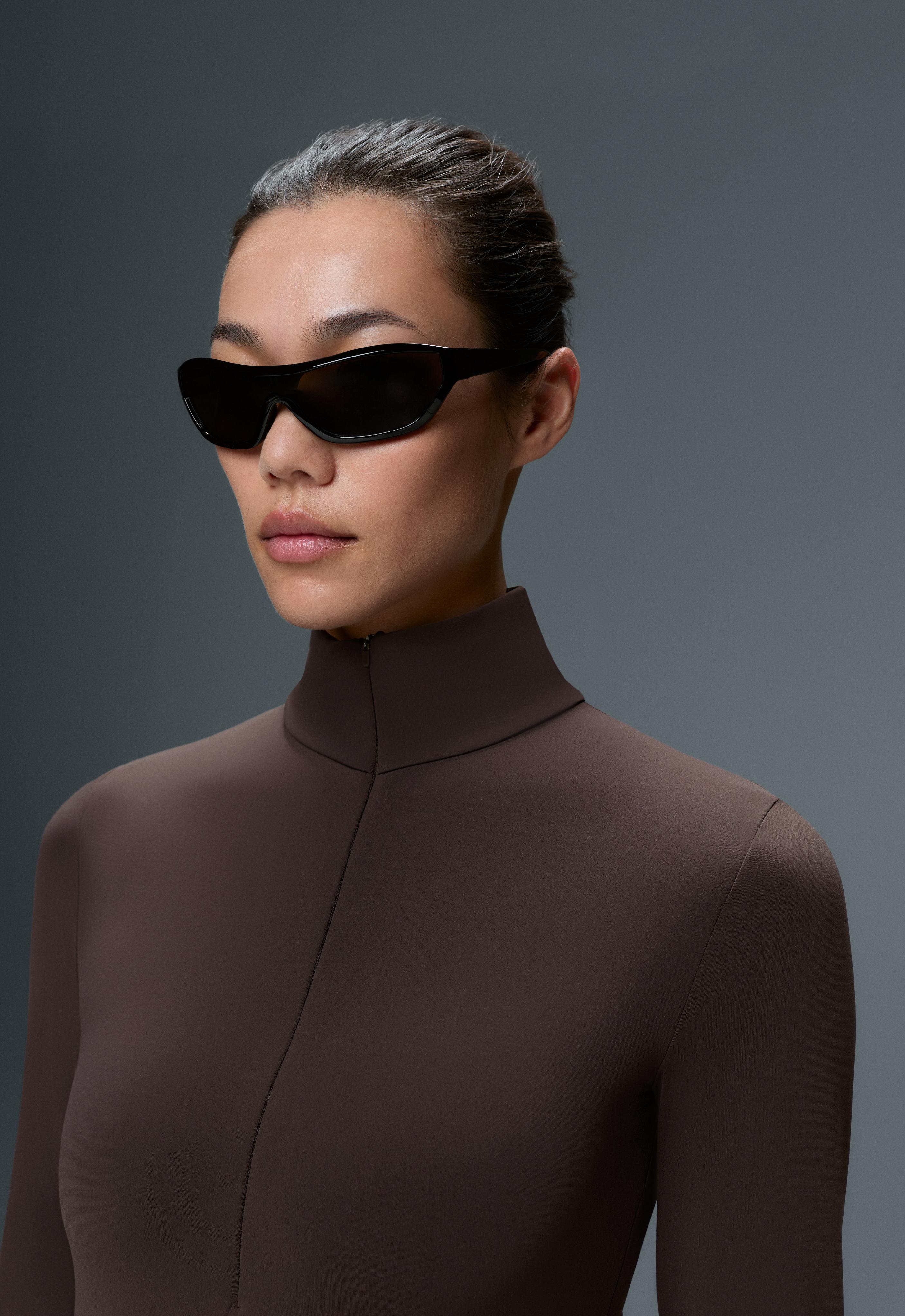 Close-up of a dark brown long-sleeve bodysuit with a high neckline and front seam, paired with black sunglasses.