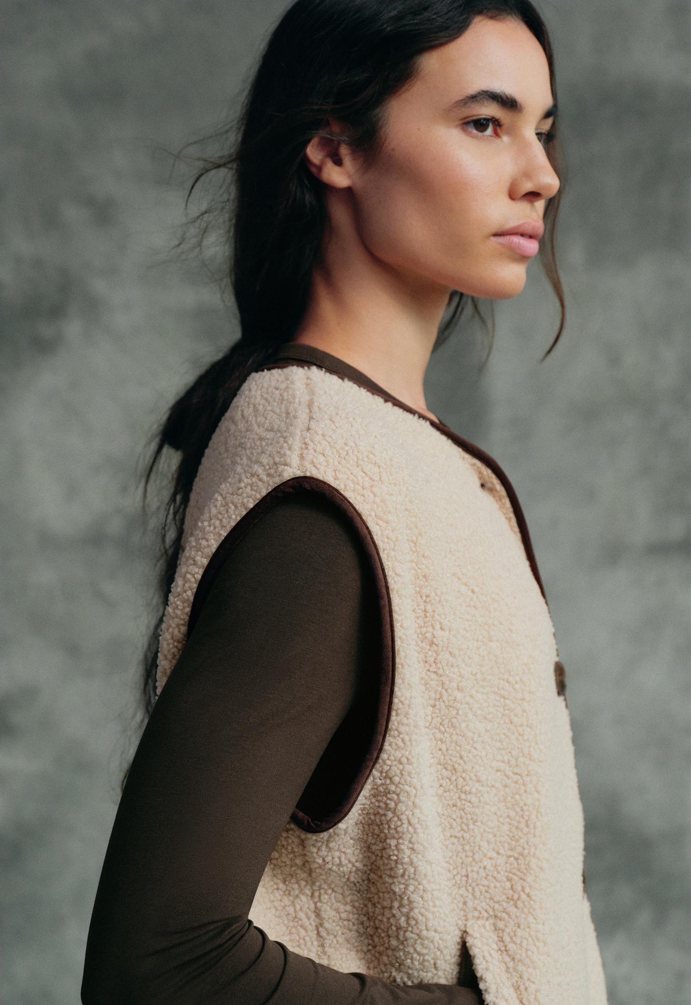 Medium shot of a cream-colored vest with contrasting trim on the armholes and neckline, a front button closure, and a side pocket, over a brown T-shirt.