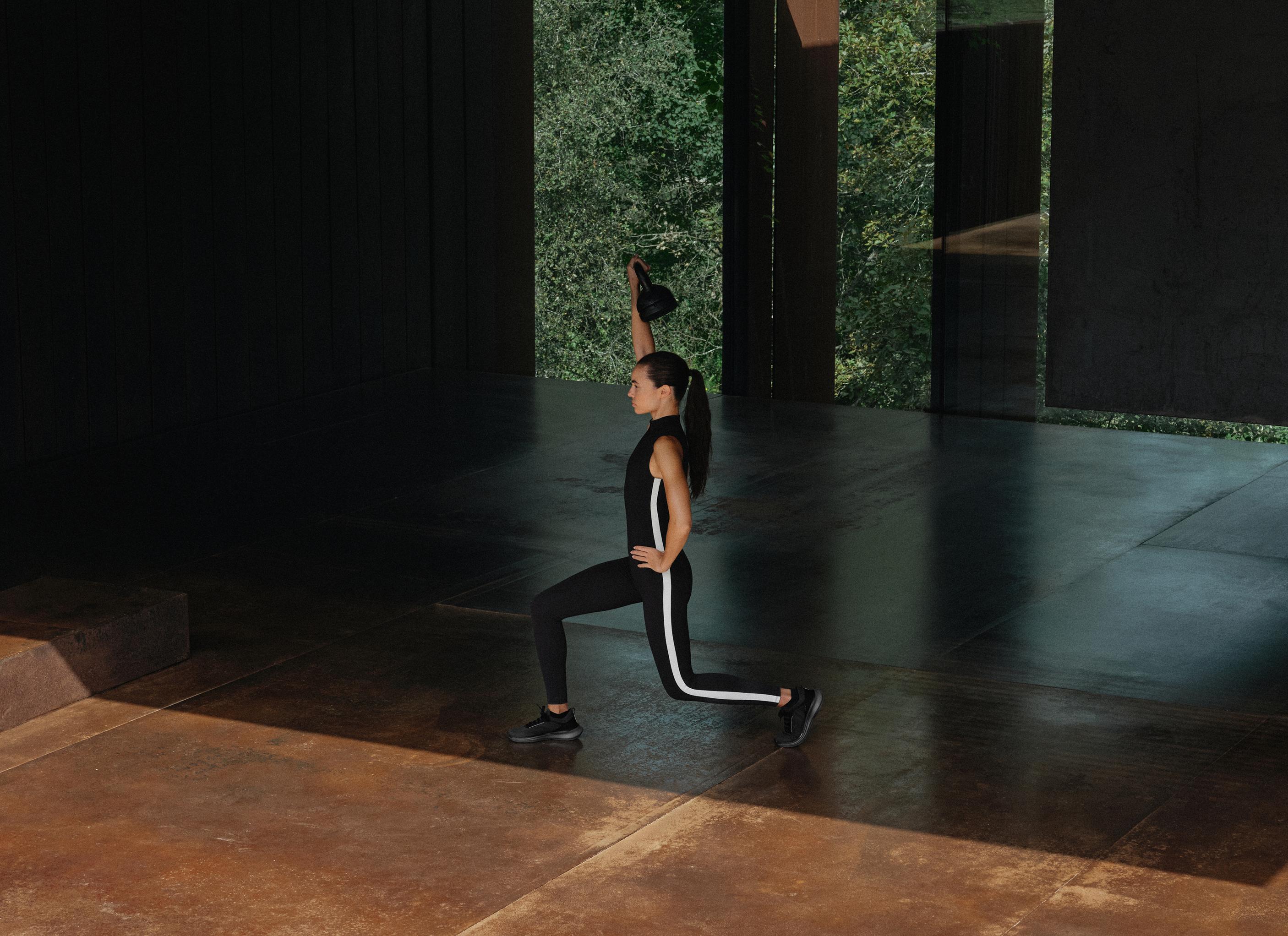 Black sleeveless full-length jumpsuit with a high neck and contrasting white side stripe, and black sneakers.
