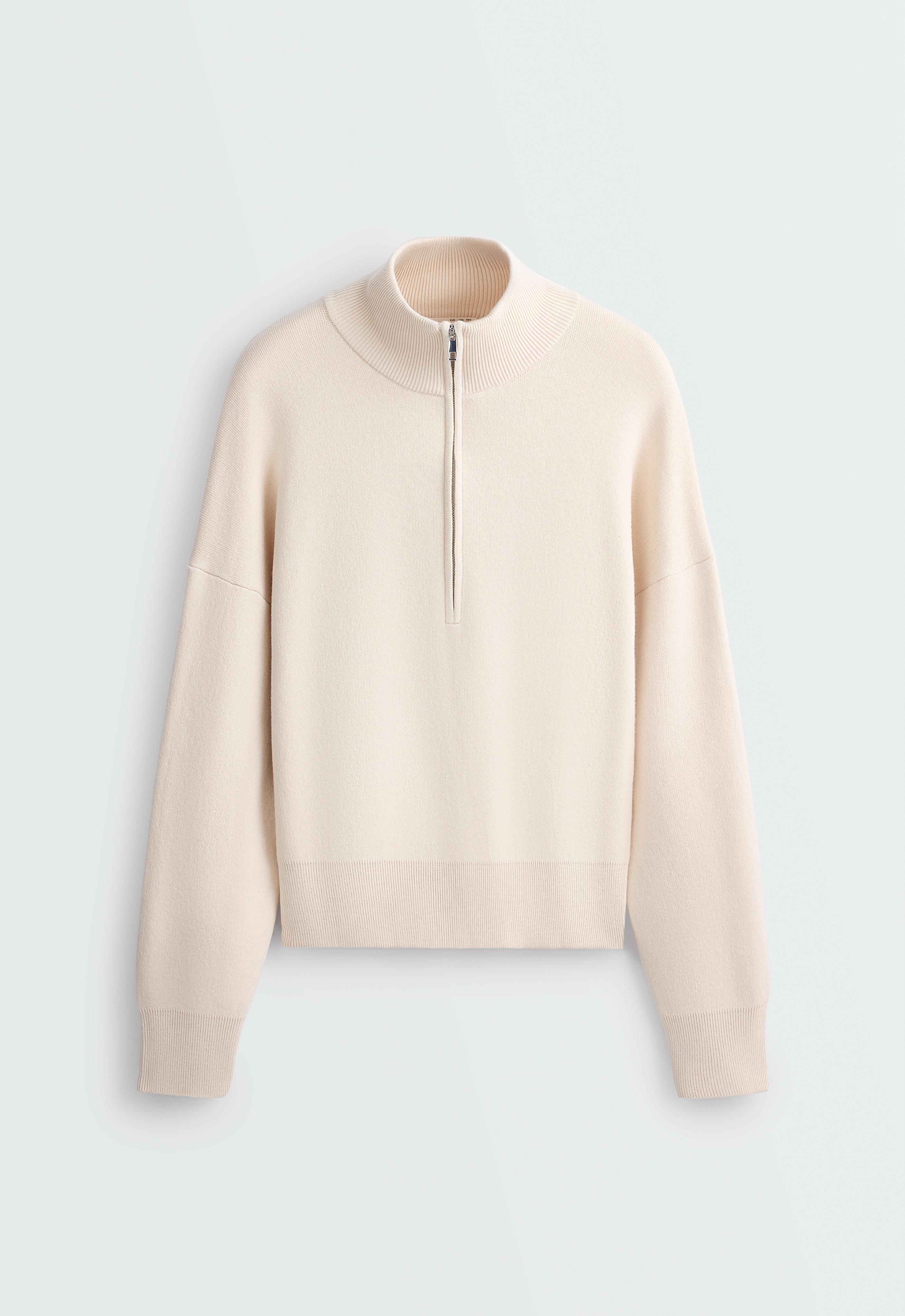 Front view of an ecru half-zip mock neck sweater. It features long sleeves and ribbed details on the collar, cuffs, and hem.