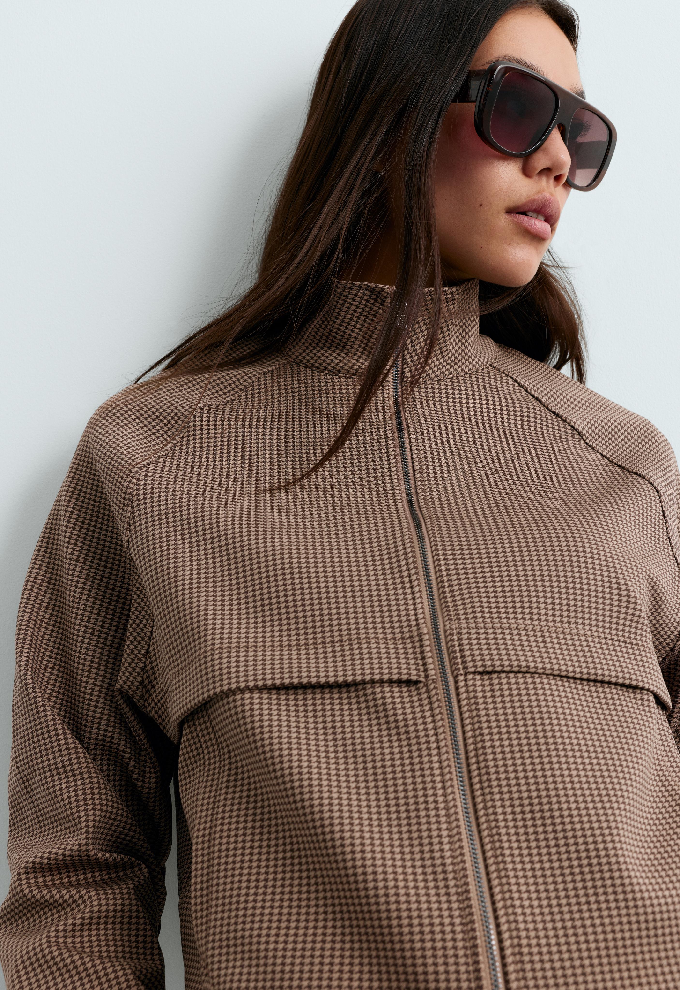 Close-up of a camel-colored jacket with a houndstooth pattern, high collar, front zipper closure, and horizontal seam across the chest.
