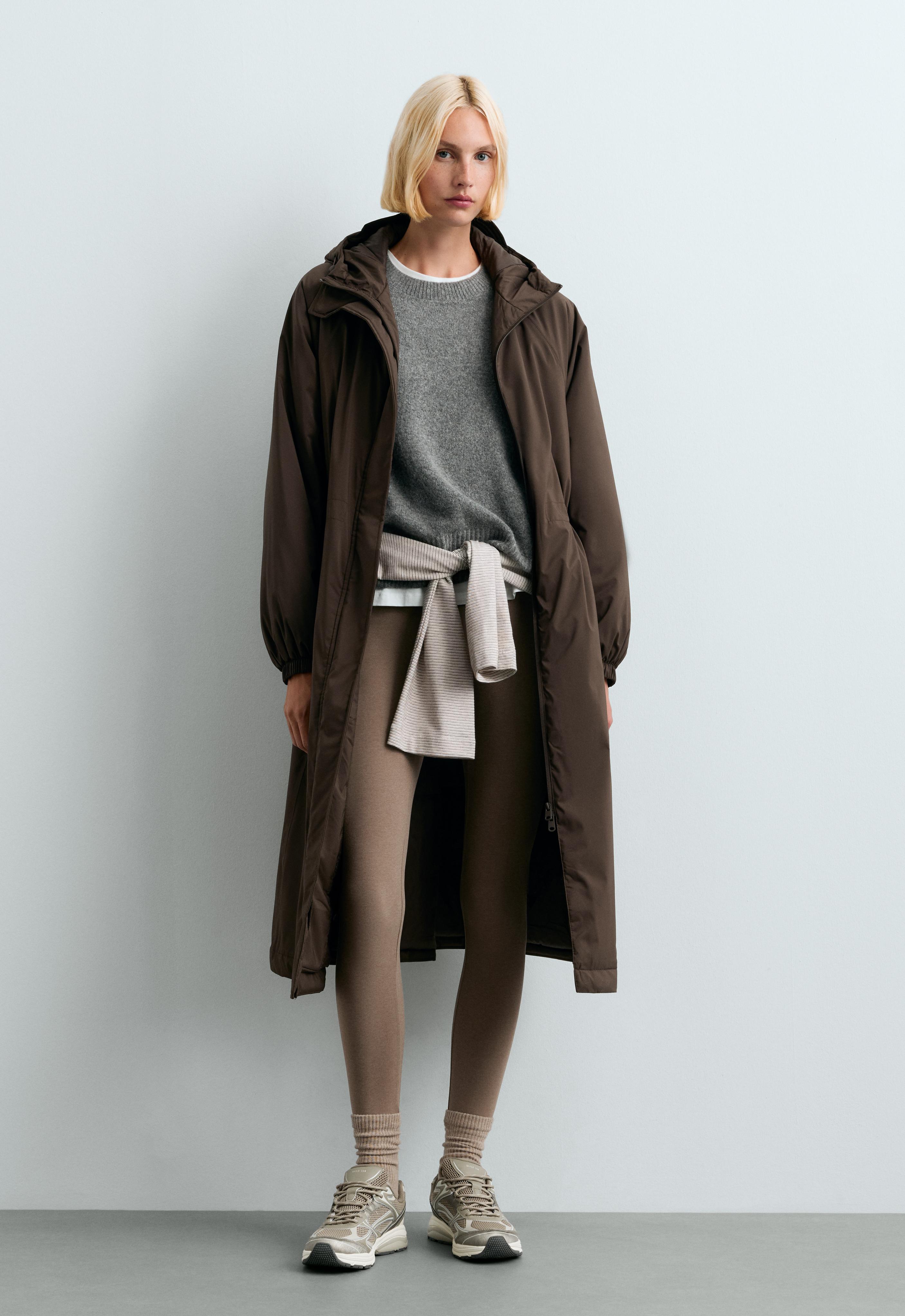Open brown long jacket with hood, gray sweater, shirt tied around the waist, brown leggings, and sneakers.