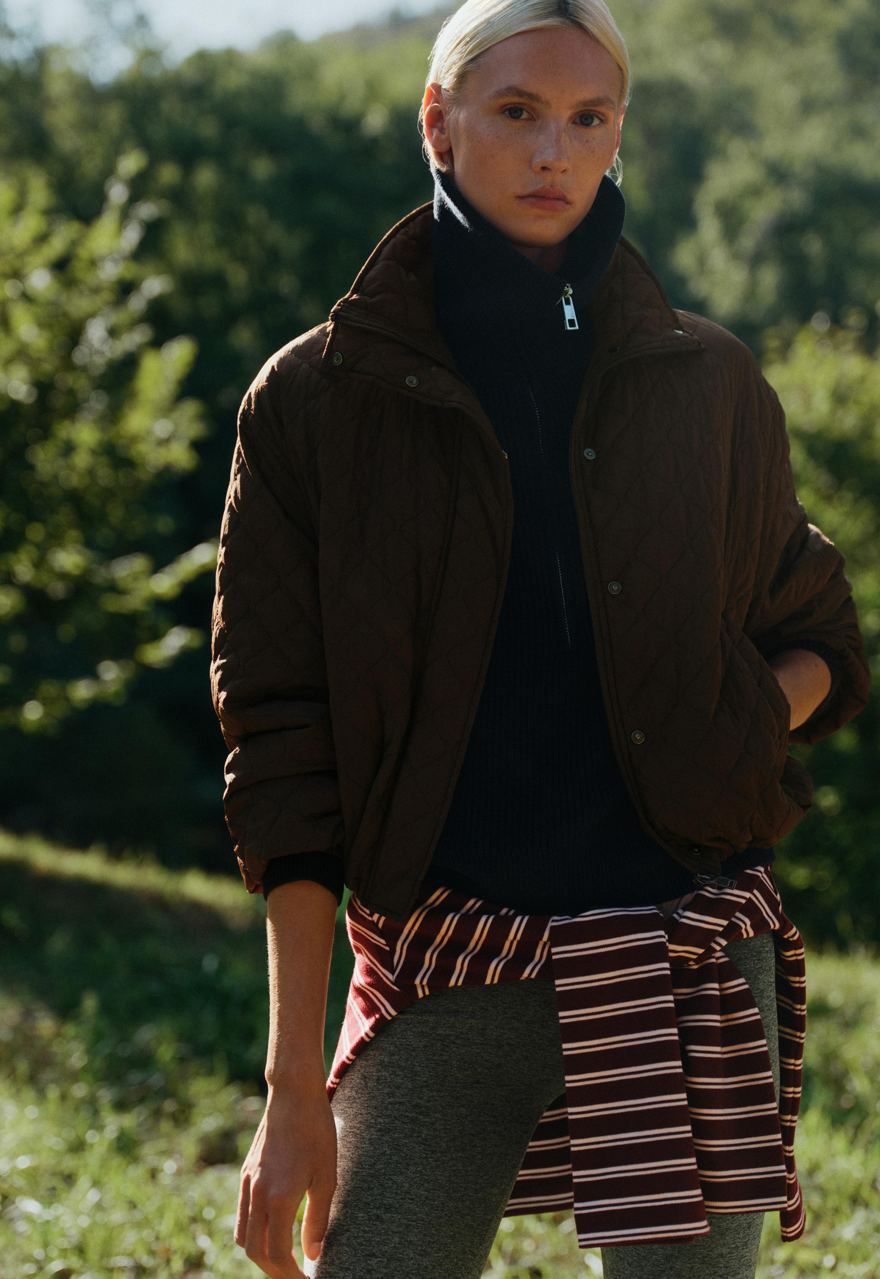 Brown quilted jacket set, navy blue mock-neck half-zip sweater, striped shirt at the waist, and gray pants.